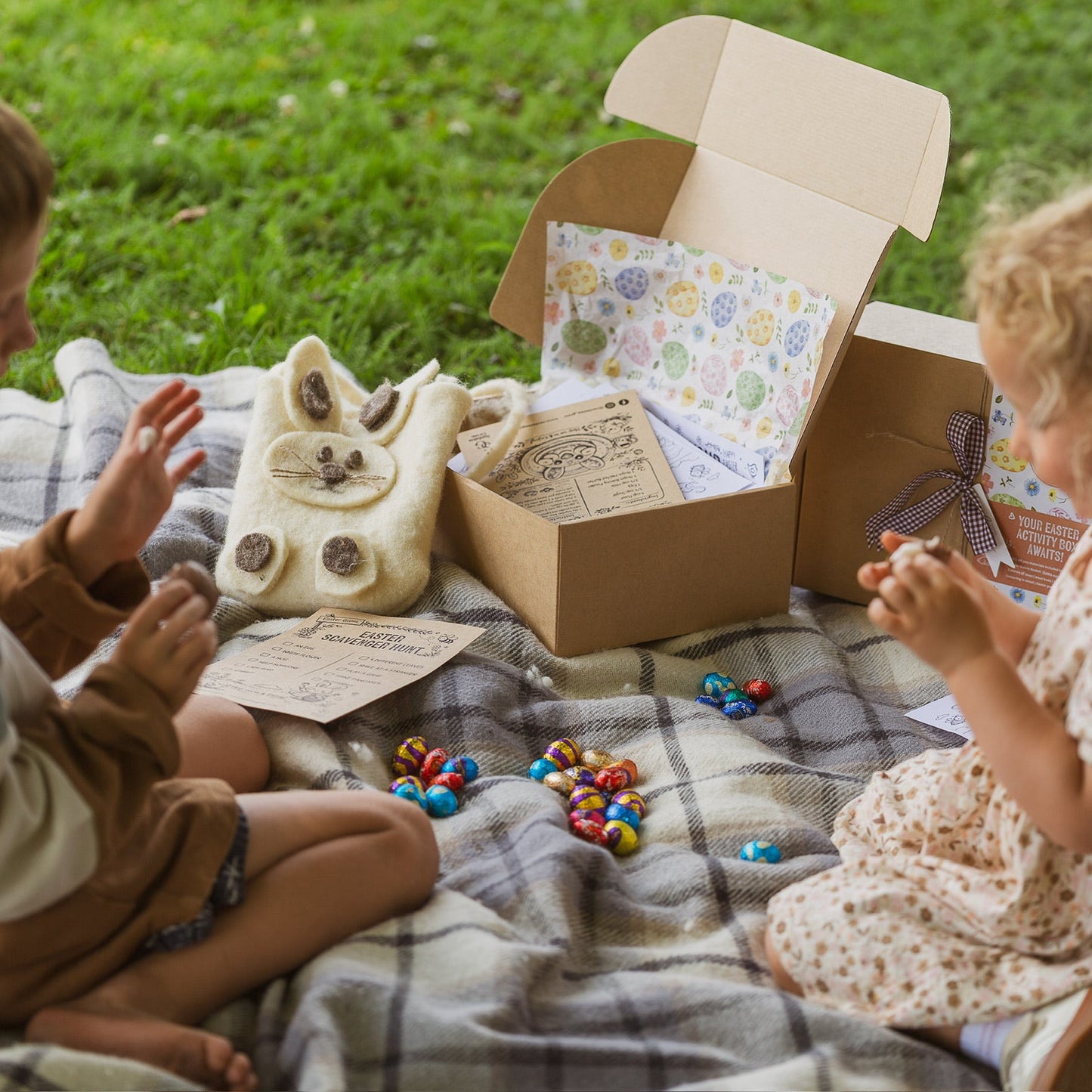 Two children sitting on a blanket outdoors with a cardboard box and toys.